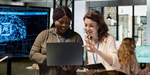 Zwei Mitarbeiterinnen arbeiten gemeinsam an einem Laptop und sprechen über den Einsatz von KI im Unternehmen.